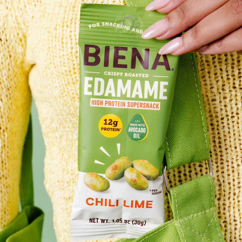 Close up picture of a person pulling a Biena Chili Lime Crispy Roasted Edamame 1.05oz bag out of their green purse